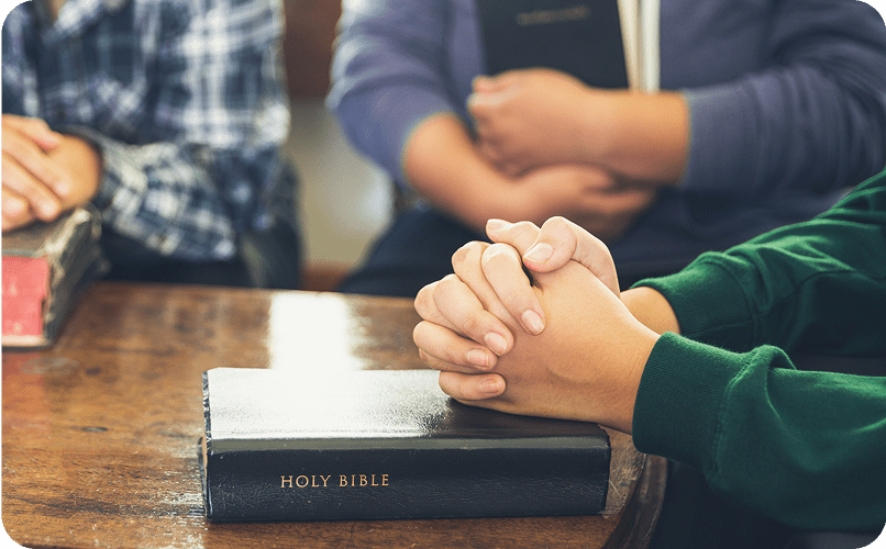 Hands praying over a Bible on table.